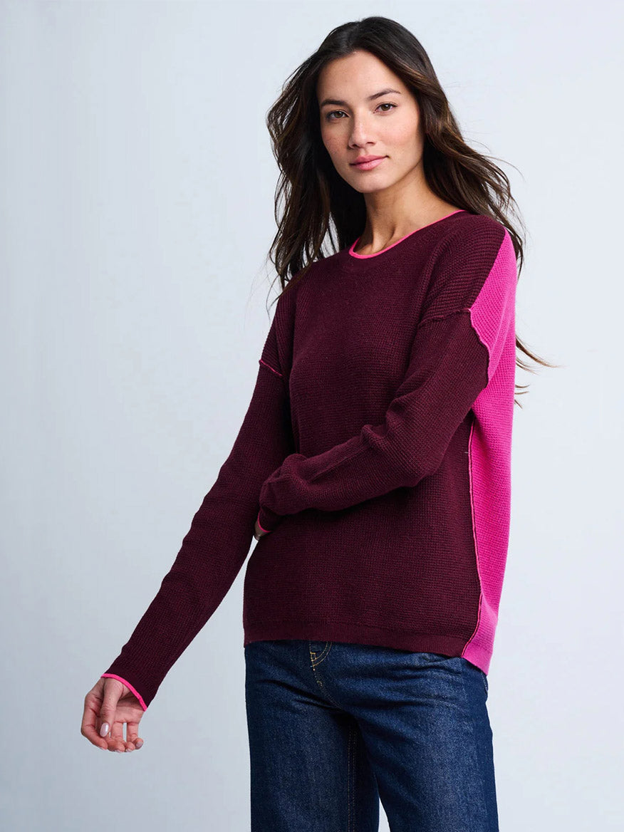 A woman wears the Lisa Todd Around Town Sweater in Wine Hot Orchid, featuring a maroon and pink colorblock with a waffle stitch texture, paired with blue jeans against a plain light background.
