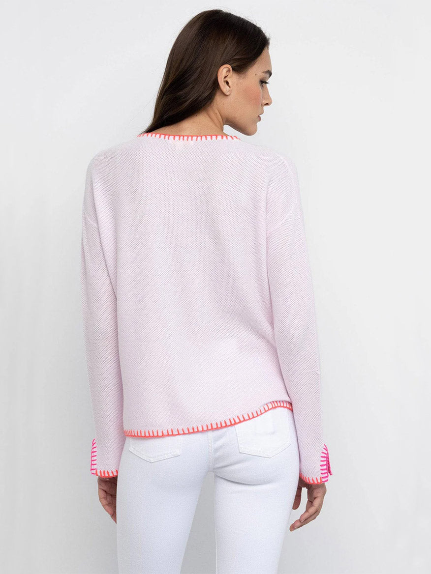 A woman stands with her back to the camera in the Lisa Todd Beach Lover V-Neck Sweater in Wisteria, featuring a relaxed fit and red trim, paired with white pants against a plain white background.