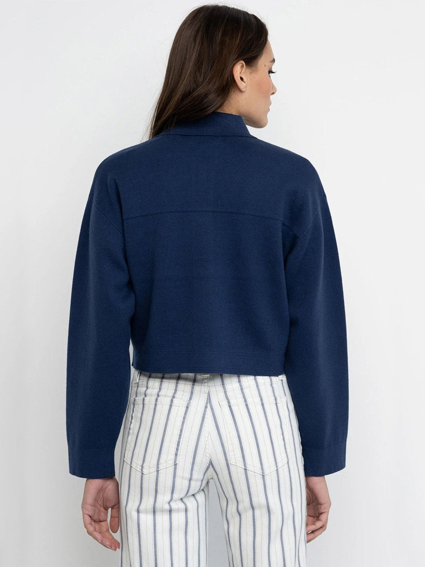 A person stands facing away, wearing the Lisa Todd Beat Goes On Jacket in Navy, a cropped denim piece with snap front closure, paired with high-waisted white pants featuring vertical blue stripes.