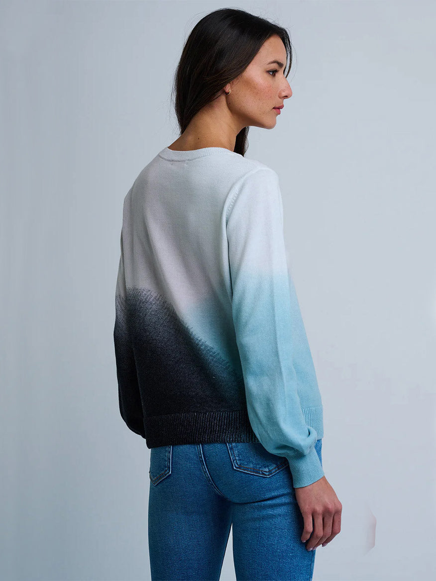 A woman stands sideways in the Lisa Todd Color Haze Sweater in Blue Combo—a 100% cotton crewneck with an ombré print from white to black to light blue—paired with blue jeans against a plain background.