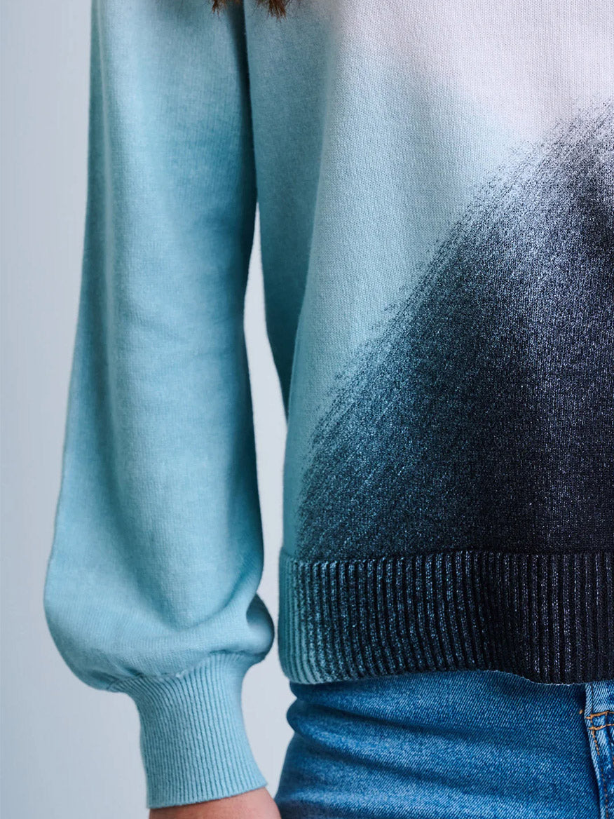 A person wears the Lisa Todd Color Haze Sweater in Blue Combo, featuring a light blue ombré crewneck with a black gradient design, paired with blue jeans and shown from shoulder to waist.