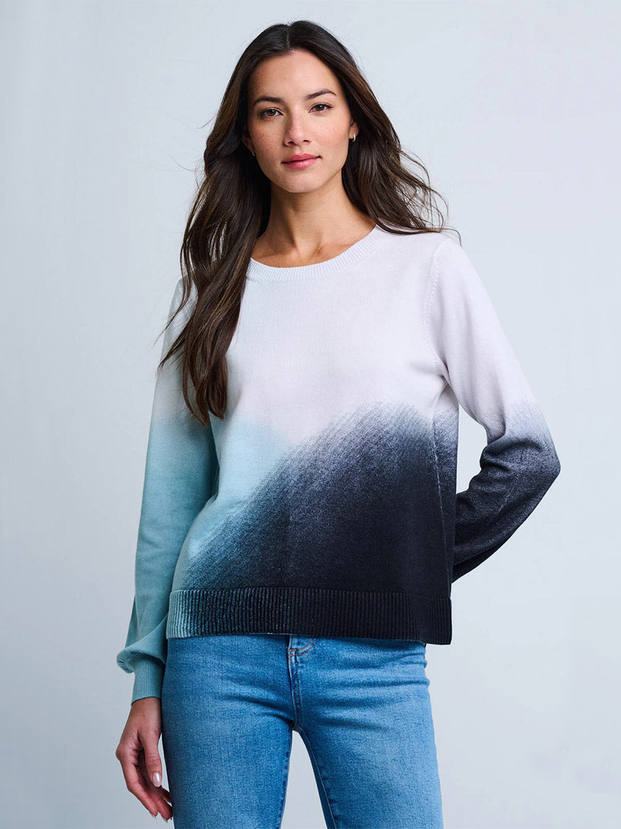 A woman poses against a plain background wearing the Lisa Todd Color Haze Sweater in Blue Combo— a long-sleeve, crewneck sweater featuring an ombré print in light blue, white, and black—paired with blue jeans.