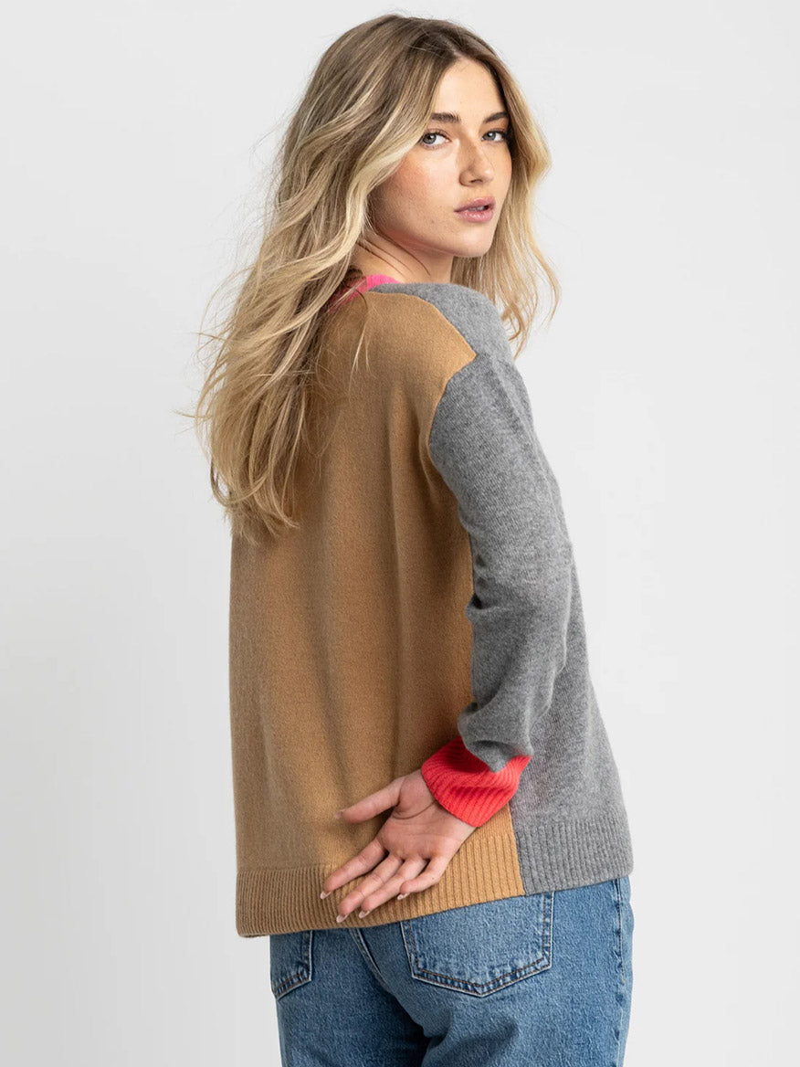 A woman with long blonde hair wears the Lisa Todd Color Wheel Sweater in Fog/Caramel, paired with blue jeans, standing slightly turned away from the camera against a light background.