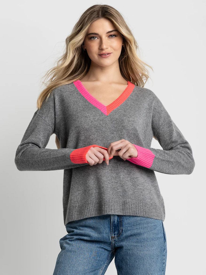 A woman wears the Lisa Todd Color Wheel Sweater in Fog/Caramel, featuring a gray colorblock design with pink and orange trim, paired with blue jeans against a plain light background.
