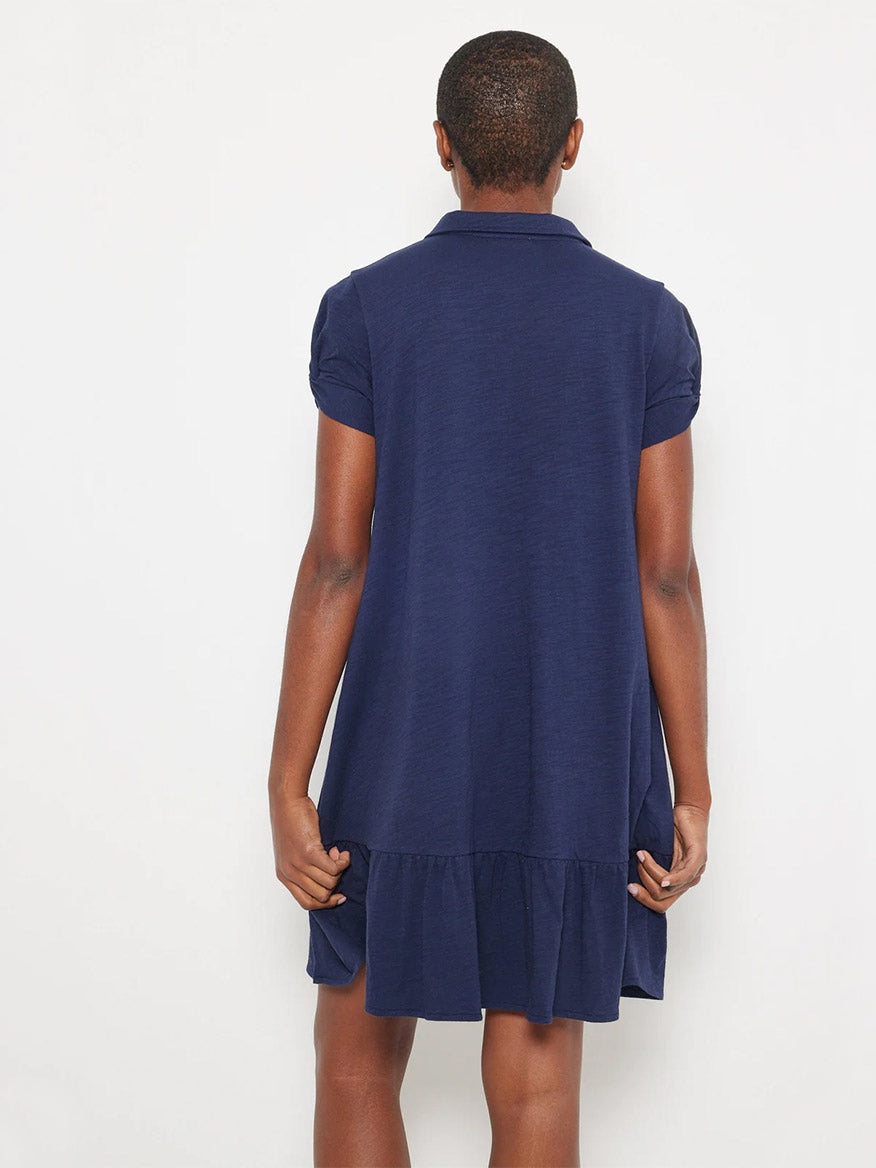 A person stands with their back to the camera, wearing the Lisa Todd Courtside Dress in Navy—a cotton knit design with ruched short sleeves and a ruffled hem—set against a plain background.