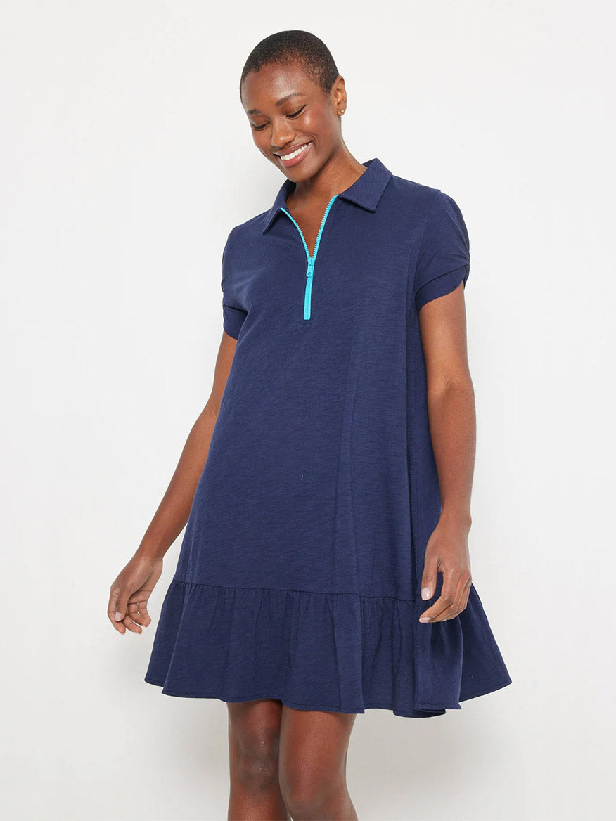 A person stands against a white background, looking down and smiling, wearing the Lisa Todd Courtside Dress in Navy, featuring ruched short sleeves, a collar, contrast zip-up closure, and a ruffled hem.
