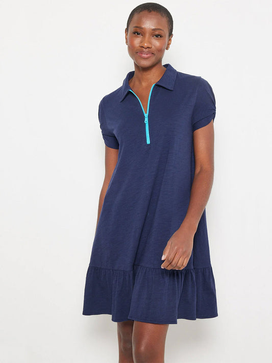 A woman wears the Lisa Todd Courtside Dress in Navy, featuring ruched short sleeves, a ruffled hem, and a bright blue contrast zip-up closure, standing against a plain light background.