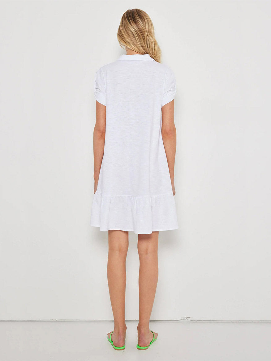 A woman stands facing a white wall, wearing the Lisa Todd Courtside Dress in White with ruched sleeves and a ruffled hem, paired with green flip-flops.