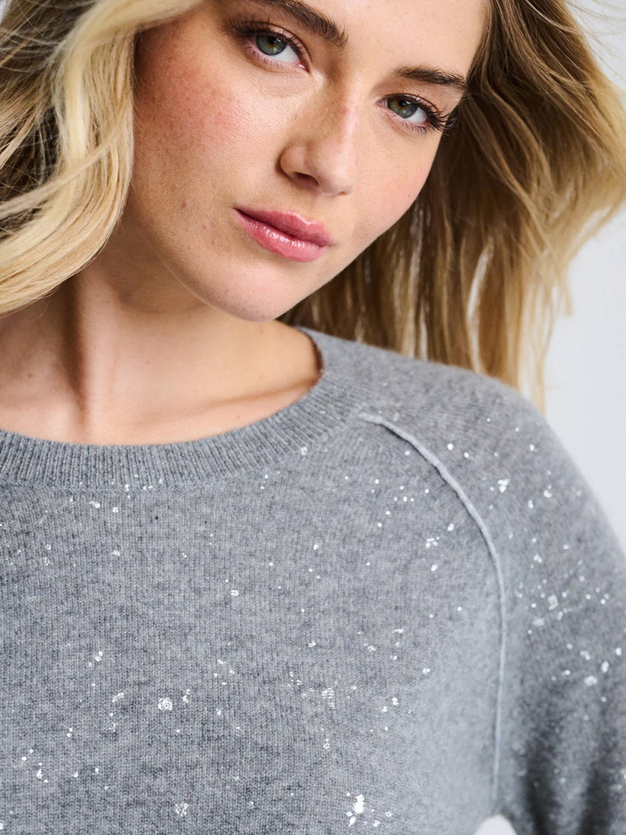 A woman with blonde hair wears the Lisa Todd Diamond Dust Sweater in Fog, a relaxed fit light grey style with white speckles, looking slightly past the camera against a plain background.