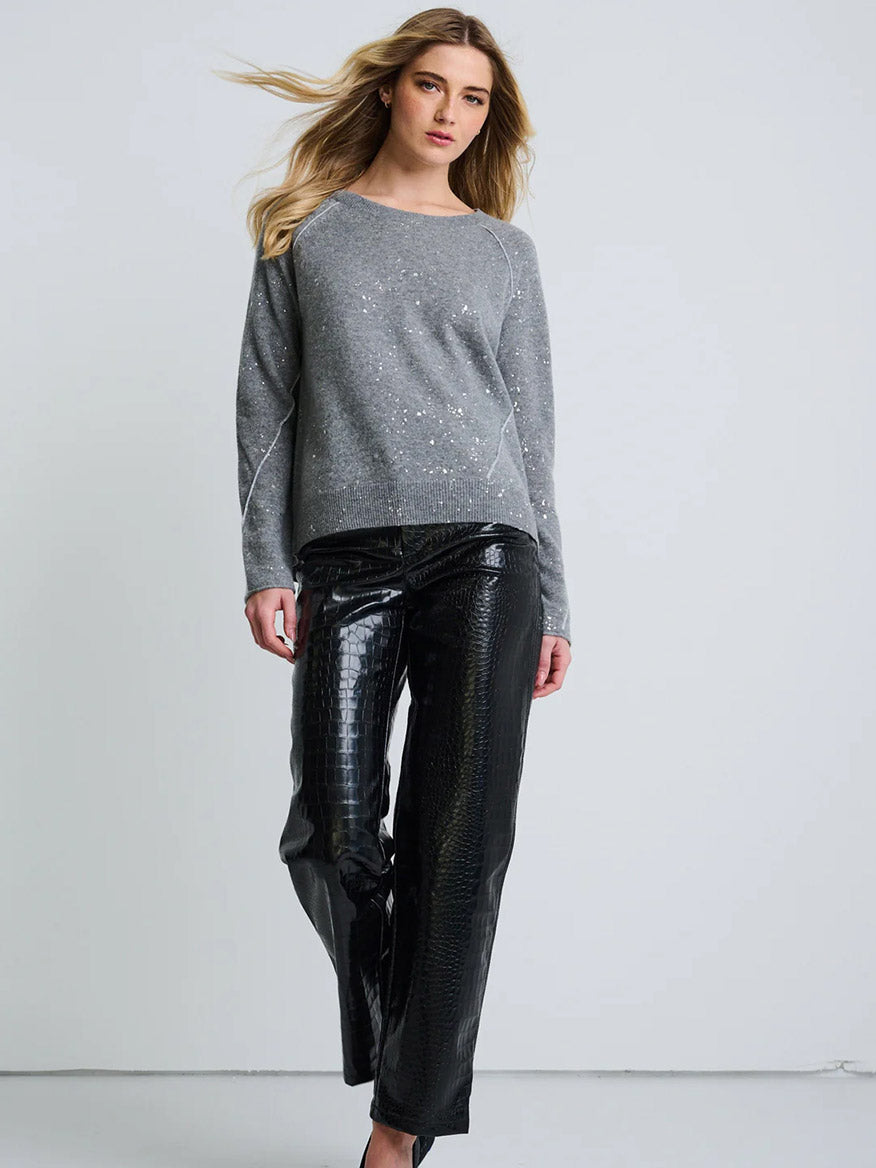 A woman stands against a plain background wearing the Lisa Todd Diamond Dust Sweater in Fog and black faux-leather pants with a textured pattern.