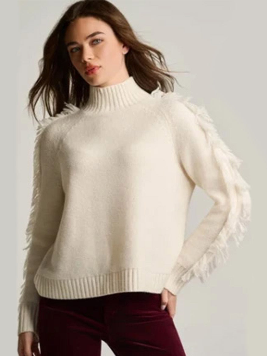 Woman wearing a Lisa Todd Fringe Benefits Sweater in Winter White—high-neck, relaxed raglan style with fringe—paired with maroon pants against a plain background.