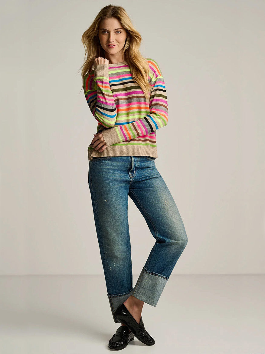 Woman poses in the Lisa Todd Hype The Stripe Sweater in Oat Multi Stripe, cuffed blue jeans, and black shoes against a plain background.