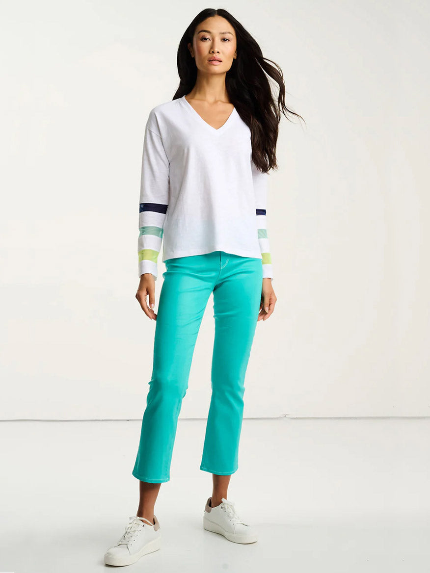 A woman stands in a white room wearing the Lisa Todd Mesh Fest Cotton Tee in White, featuring colored stripes on the sleeves, paired with teal cropped pants and white sneakers.