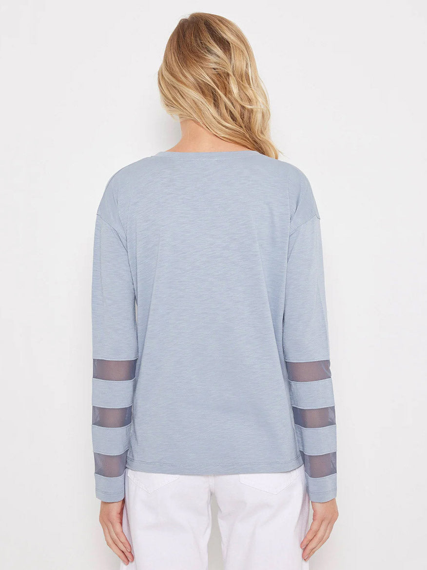A woman with long blonde hair, seen from behind, wears the Lisa Todd Mesh Fest Top in Denim—a light blue V-neck with sheer mesh striped sleeves—paired with white pants.