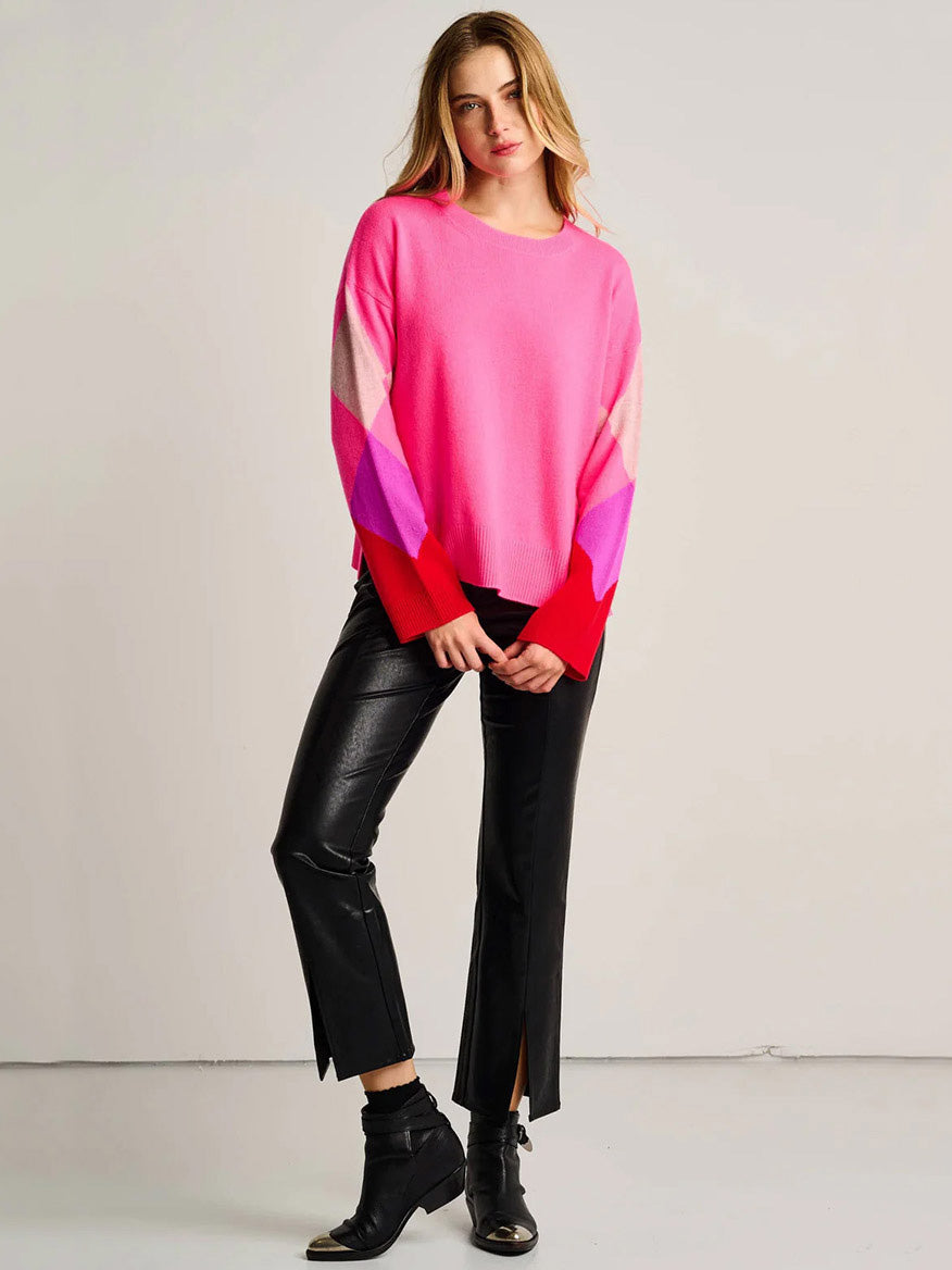 A woman stands against a plain background wearing the Lisa Todd Power Surge Sweater in Neon Pink, paired with relaxed fit black leather pants with front slits and black ankle boots.