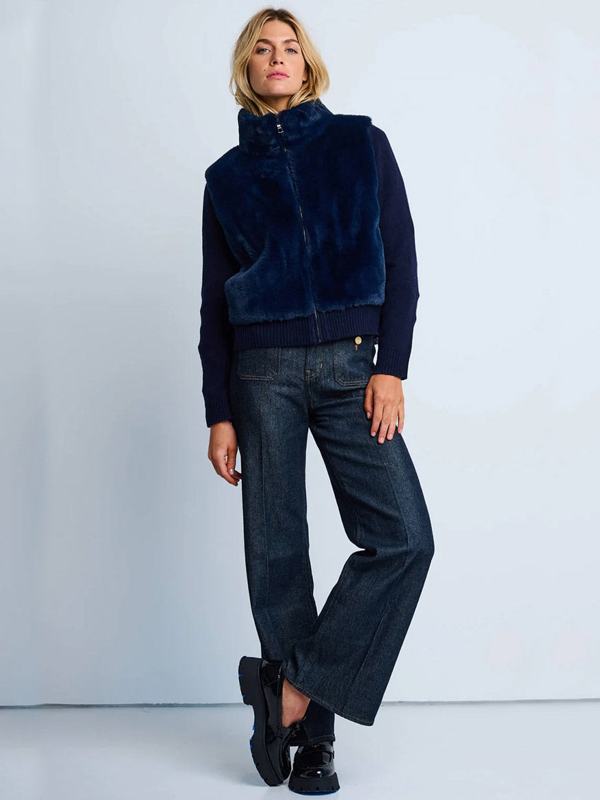 A person stands against a plain background wearing the Lisa Todd Teddy Faux Fur Knit Jacket in Navy, a dark sweater, wide-leg jeans, and black shoes.