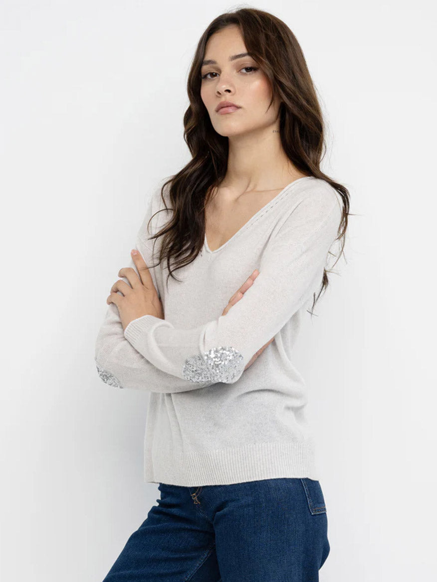 Woman wearing the Lisa Todd Truth or Dare Reversible Sweater in White, a relaxed fit V-neck with sequined elbow patches, paired with blue jeans and standing with arms crossed against a plain white background.