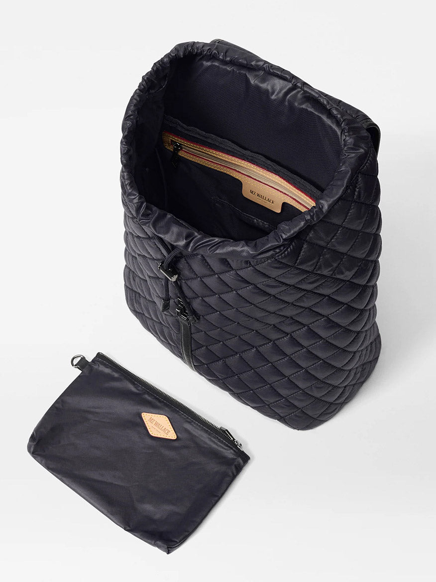 The MZ Wallace Astor Backpack in Black Oxford is shown open with gunmetal hardware, an inner zip pocket visible, and a matching zippered pouch placed beside it.