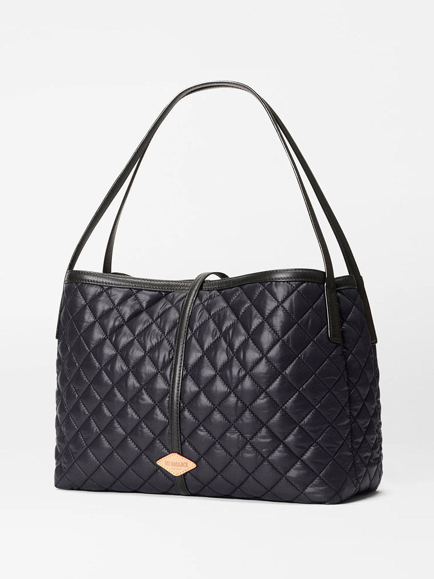 The MZ Wallace Astor East West Tote in Black Oxford features black quilted Italian leather, double shoulder straps, gunmetal hardware, and a gold emblem on the front, shown against a plain white background.