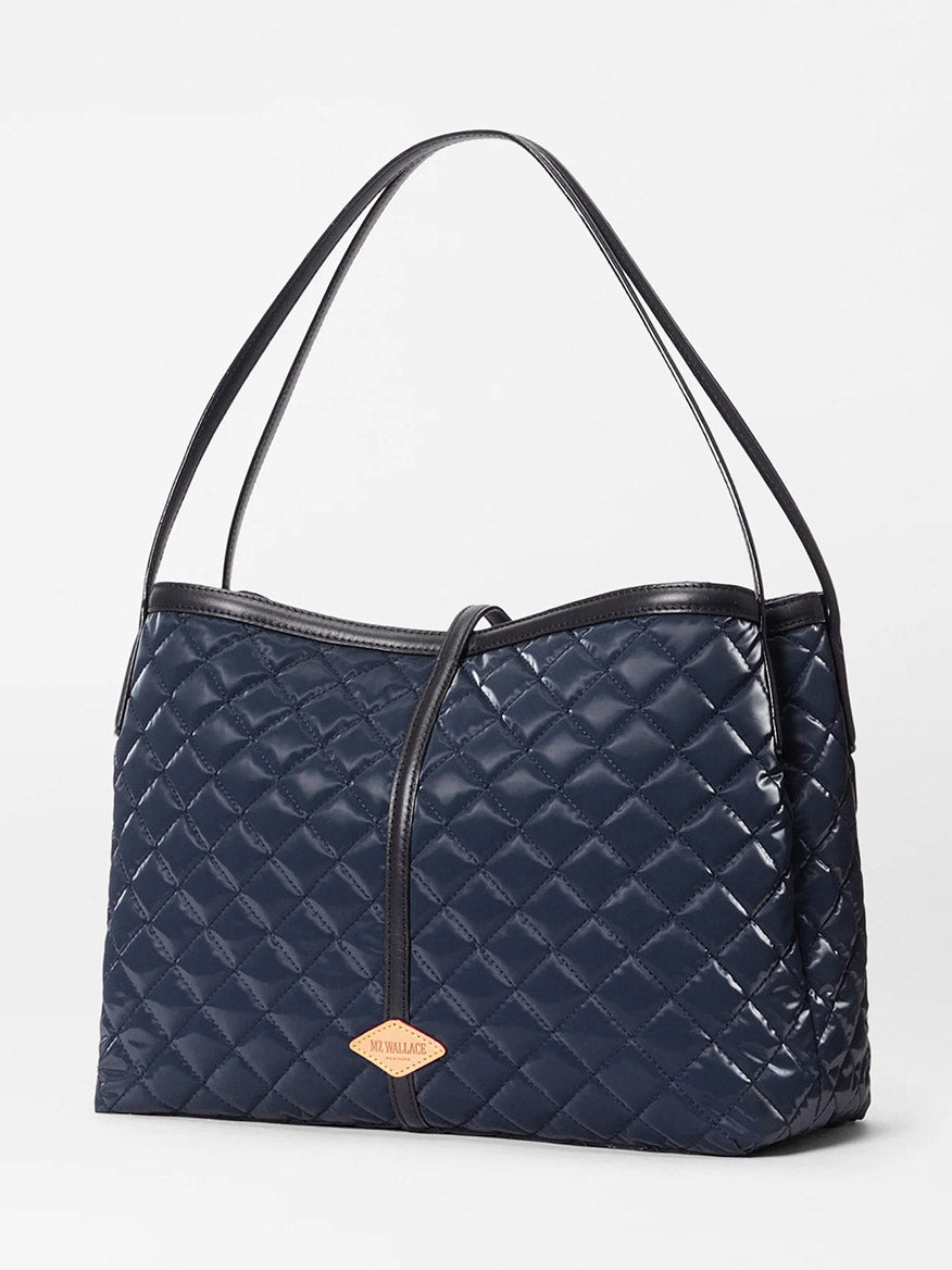 The MZ Wallace Astor East West Tote in Ink Liquid Oxford features a navy blue quilted design, black Italian leather handles, gunmetal hardware, and a tan leather tag, shown against a plain white background.
