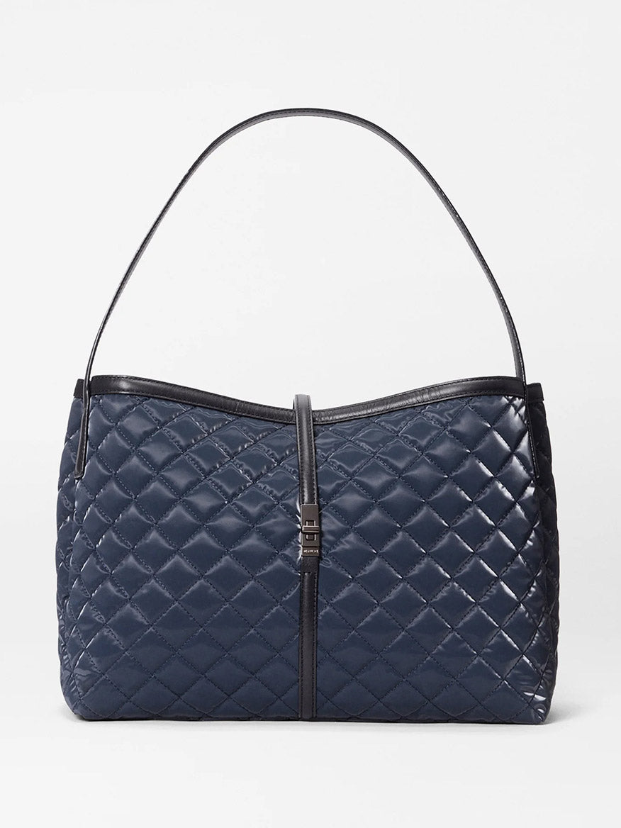 The MZ Wallace Astor East West Tote in Ink Liquid Oxford is a navy blue quilted bag with a single black Italian leather shoulder strap and a central vertical black leather accent finished with a metal clasp.