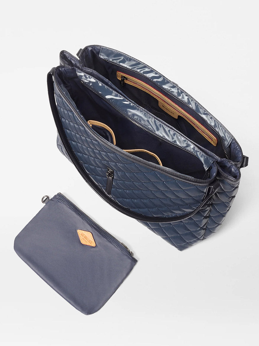 The MZ Wallace Astor East West Tote in Ink Liquid Oxford features a quilted navy design, open top with turnlock strap closure, interior pocket for glasses and valuables, and includes a coordinating zippered pouch. Versatile enough for use as a laptop bag.