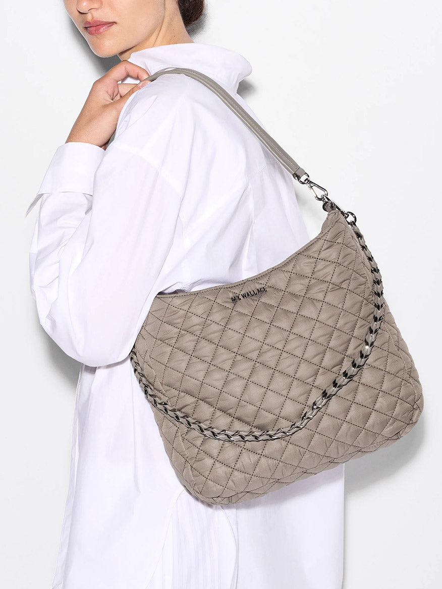 A woman in a white shirt carries the spacious MZ Wallace Crosby Hobo in Morning Dove Oxford, a quilted beige shoulder bag made from recycled nylon with Italian leather accents and a braided strap.