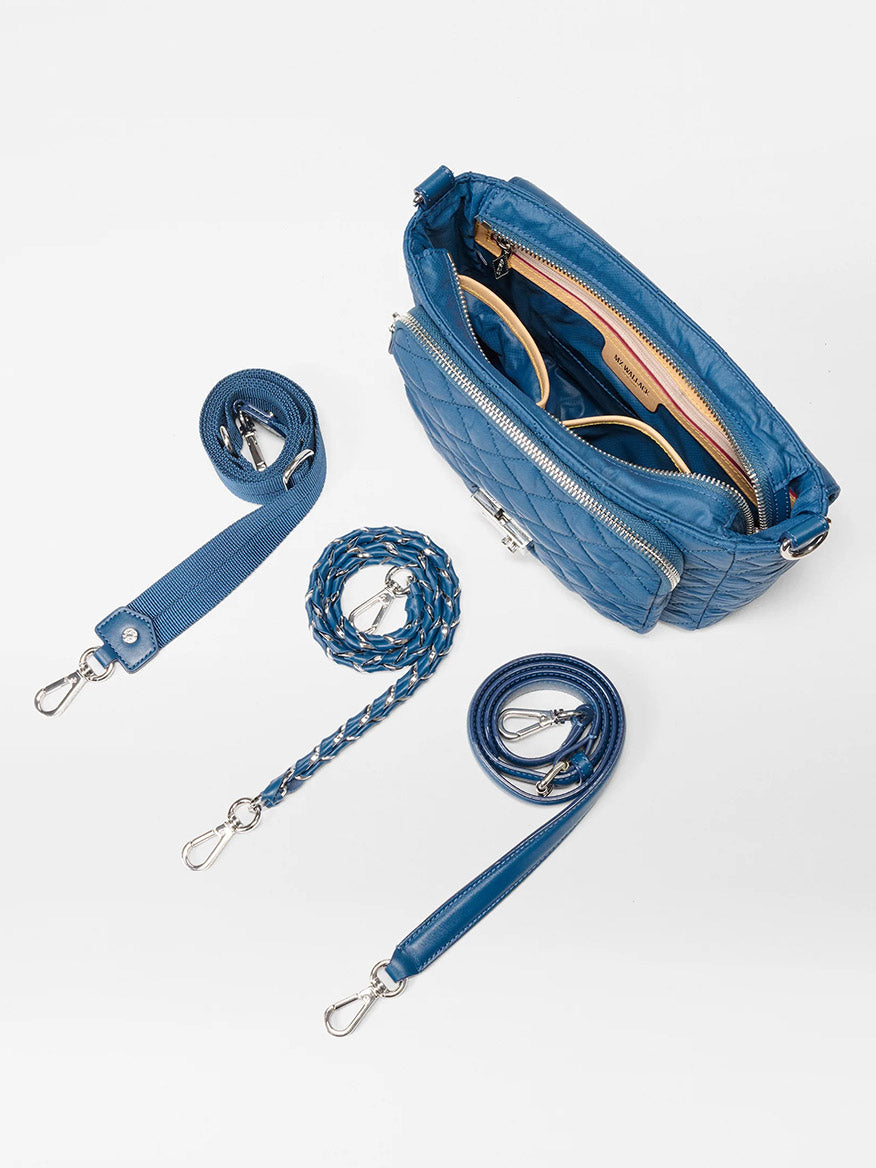 The MZ Wallace Crosby Lock Medium Crossbody in French Blue Oxford, featuring gold zippers, is shown partially open with sunglasses and a wallet inside. Three detachable blue straps are displayed beside the bag.