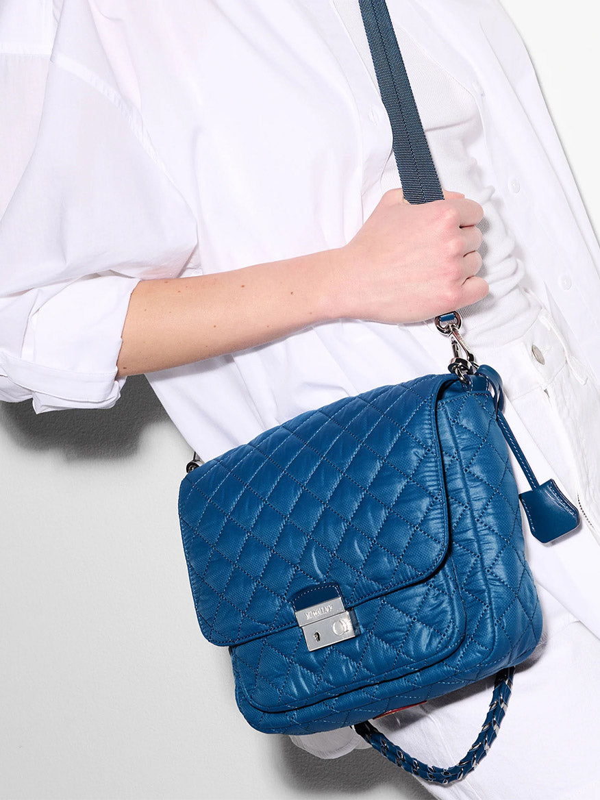 A person in a white shirt holds the MZ Wallace Crosby Lock Medium Crossbody in French Blue Oxford, a quilted designer bag with a silver clasp and adjustable shoulder strap.