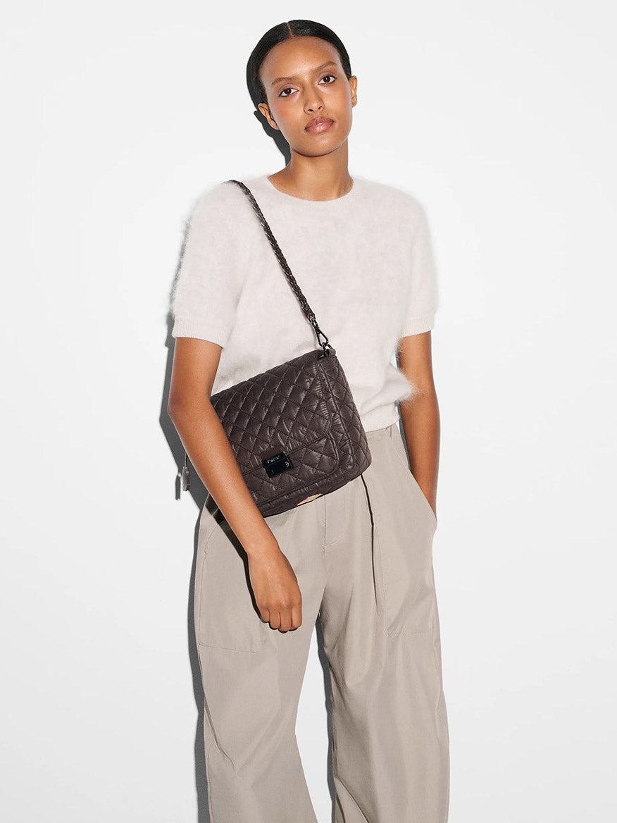 A person in a white short-sleeve sweater and beige wide-leg pants stands against a white wall, carrying the MZ Wallace Crosby Lock Medium Crossbody in Espresso Oxford over their shoulder.