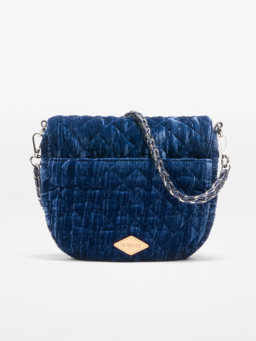 The MZ Wallace Crosby Lock Saddle in Midnight Velvet is a small quilted navy blue velvet crossbody bag with a detachable chain strap and a tan logo patch on the front.