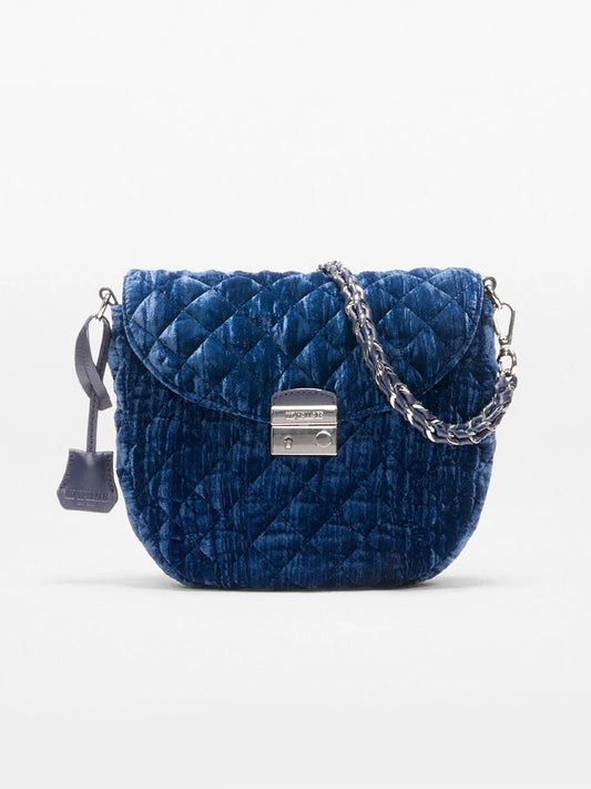 The MZ Wallace Crosby Lock Saddle in Midnight Velvet is a blue quilted velvet crossbody bag featuring a silver chain strap, metal clasp closure, and a small leather tag, combining elegance with functionality.