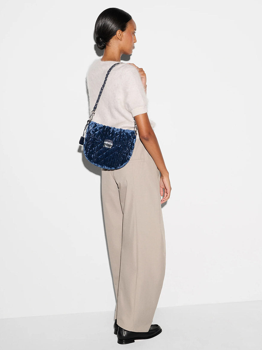 A woman stands sideways against a white background, wearing a short-sleeve white sweater, beige wide-leg pants, black shoes, and carrying the MZ Wallace Crosby Lock Saddle in Midnight Velvet with a detachable strap.