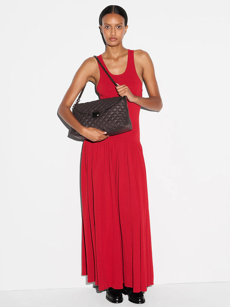 A woman in a sleeveless red maxi dress stands against a white background, holding the MZ Wallace Crosby Lock Shoulder in Espresso Oxford, paired with black shoes.