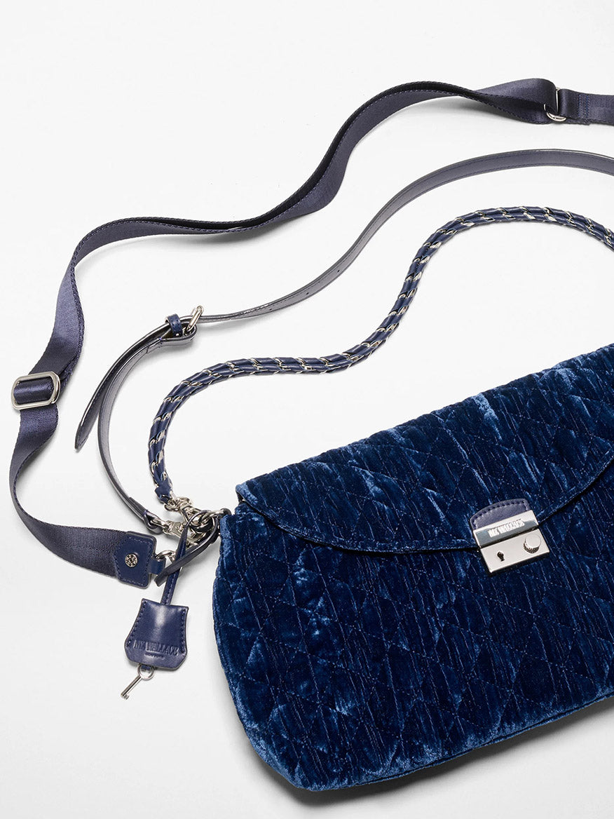 The MZ Wallace Crosby Lock Shoulder in Midnight Velvet features quilted blue velvet, Italian leather trim, a silver clasp, chain strap, and key charm, all shown on a white surface.