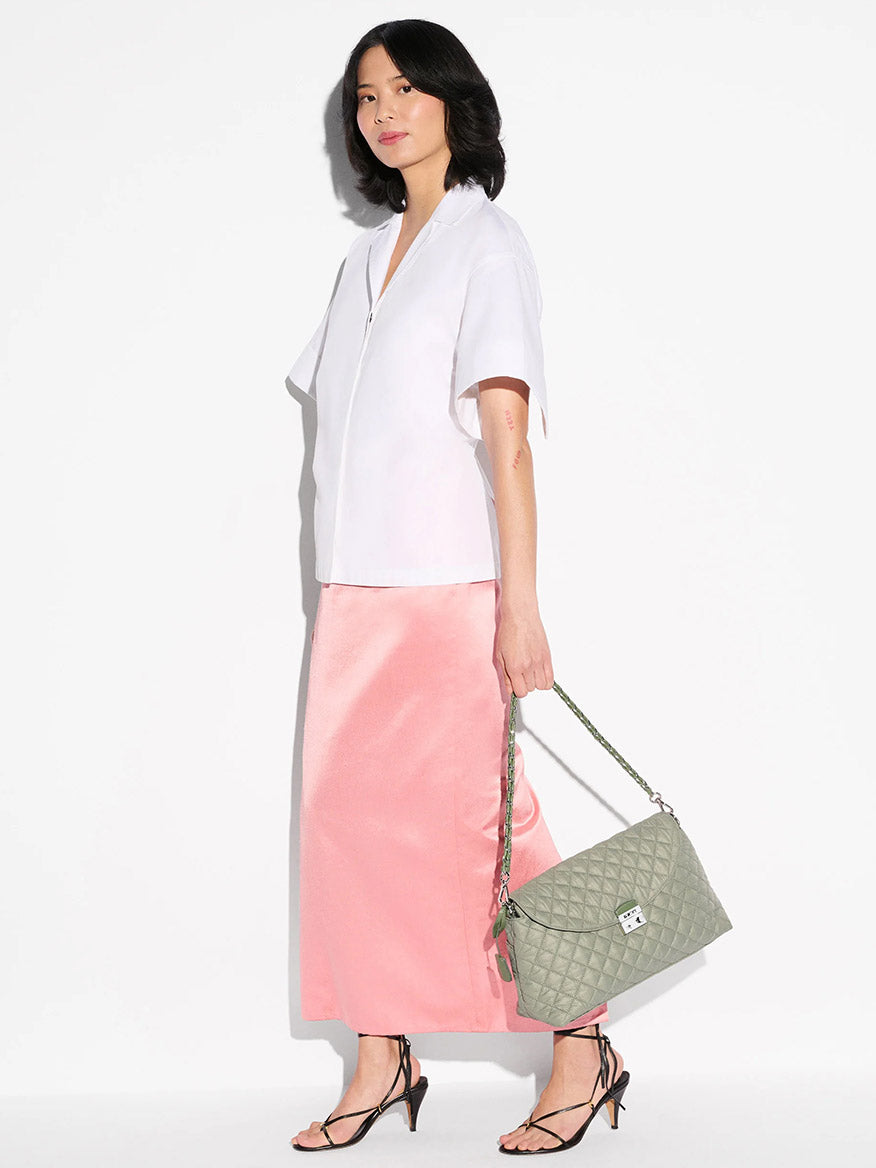 Woman stands against a white background in a short-sleeve white blouse, pink satin skirt, black strappy heels, carrying the MZ Wallace Crosby Lock Shoulder in Agave Green Oxford with Italian leather trim.