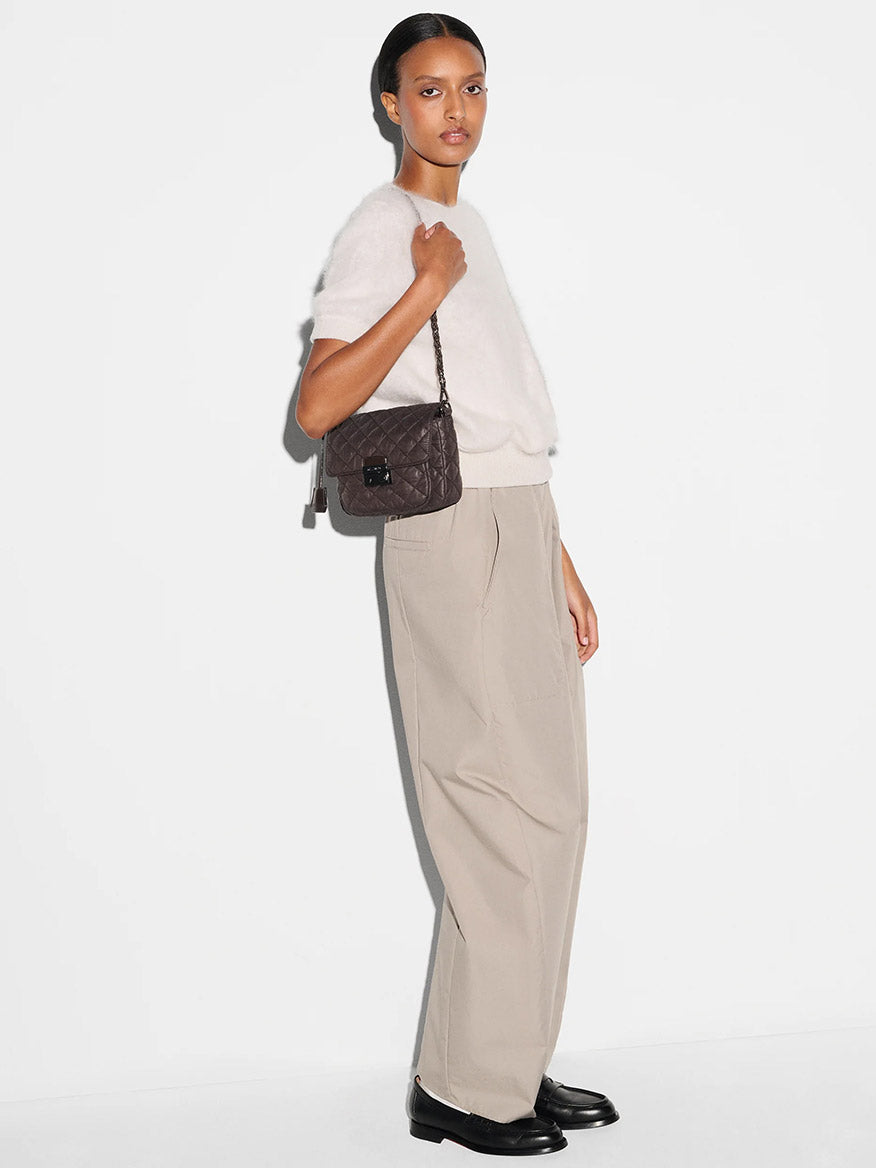 A woman stands against a white background wearing a white short-sleeve sweater, beige wide-leg trousers, black shoes, and carrying the MZ Wallace Crosby Lock Small Crossbody in Espresso Oxford.