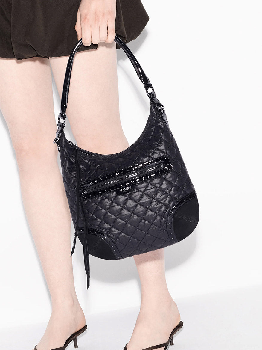 A person in black shorts and heeled sandals holds the MZ Wallace Multi Stud Crosby Sienna in Black Stud & Suede, an Oxford bag with Italian leather trim, front zipper, and shoulder strap.