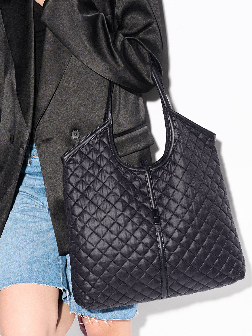 A person in a black blazer and denim skirt holds the MZ Wallace Large Astor Tote in Black Oxford, featuring Italian leather top handles.
