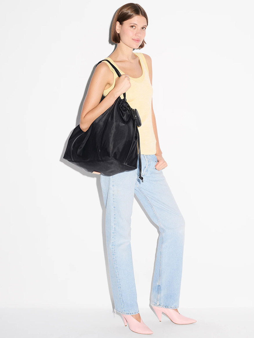 Woman stands against a white background, wearing a yellow tank top, light blue jeans, and pink heels, carrying the MZ Wallace Large Waverly Tote in Black Bedford with Italian leather handles that fits a 15" laptop over her shoulder.