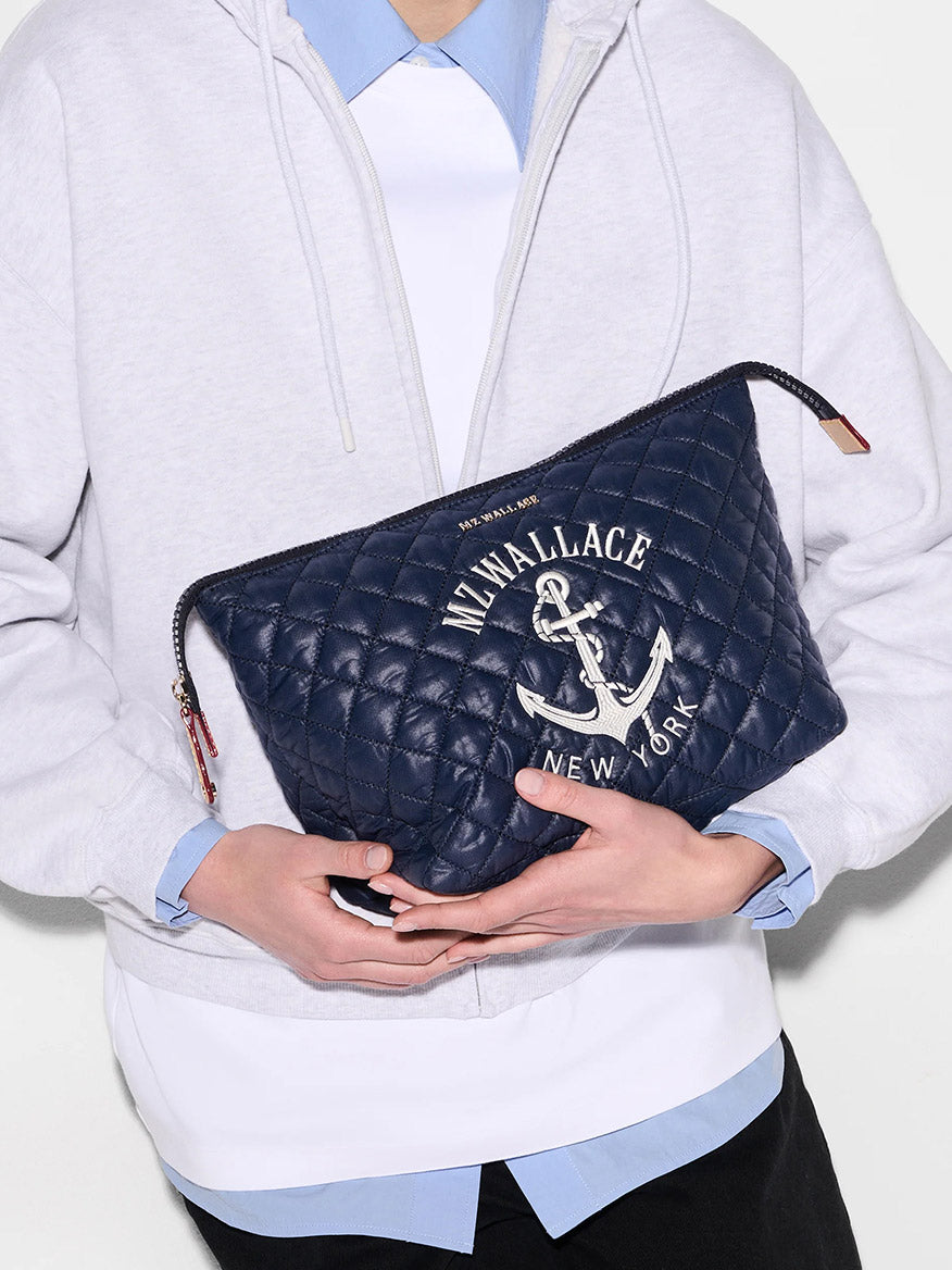 A person in a light gray hoodie and blue shirt holds the MZ Wallace Large Zoey Cosmetic in Anchor Oxford, a quilted navy makeup pouch with an anchor print that makes a stylish travel accessory.