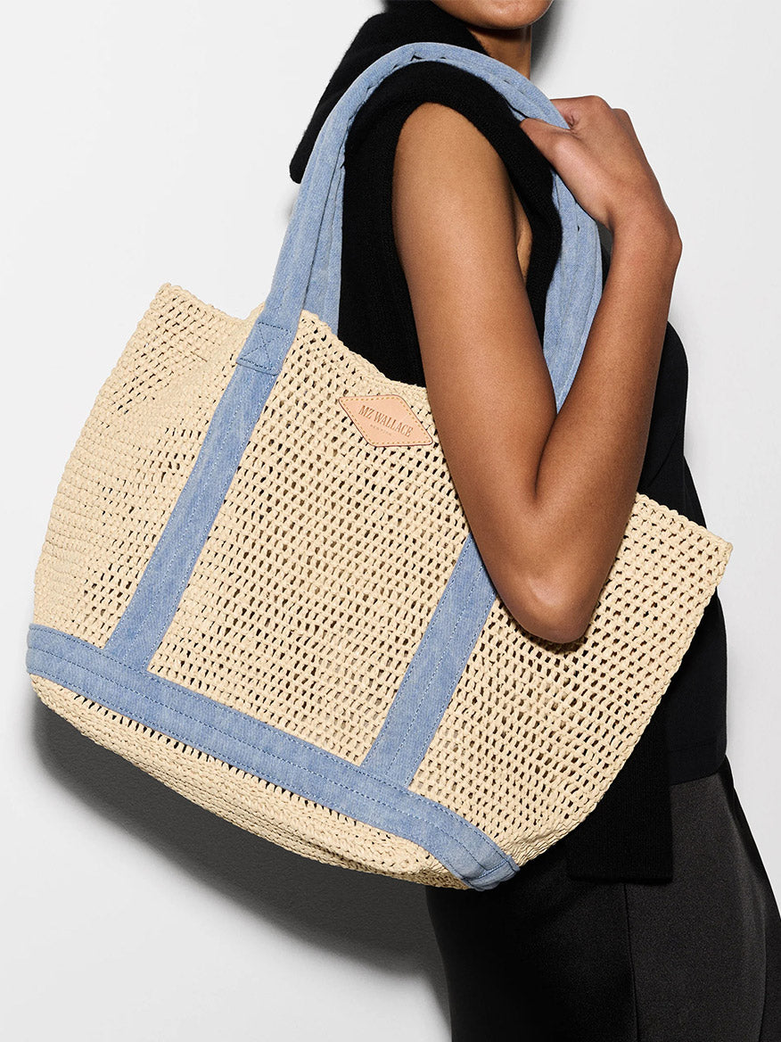 A person holds the MZ Wallace Medium Crochet Tote in Crochet/True Light Denim against a white background, showcasing its unique crochet texture and flair with blue denim straps.