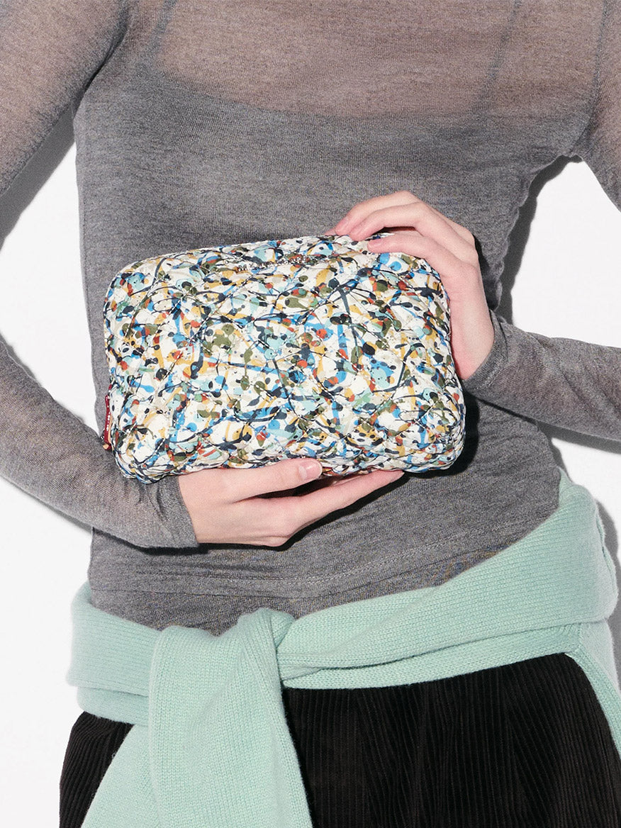 Person wearing a sheer grey top and mint sweater holds the MZ Wallace Medium Double Zip Cosmetic in Club Paint Oxford—a quilted pouch with double zip closure—in front of their torso.