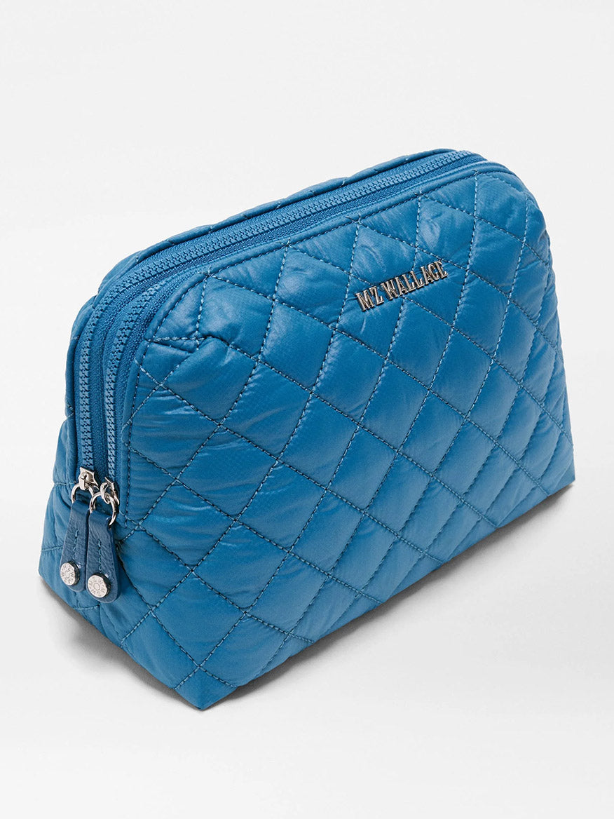 The MZ Wallace Medium Double Zip Cosmetic in Atlantic Blue Oxford stands upright on a white background, showcasing its quilted design, silver zipper pulls, and a small metallic logo on the front. Crafted from recycled nylon, it’s ideal for organizing your travel essentials.