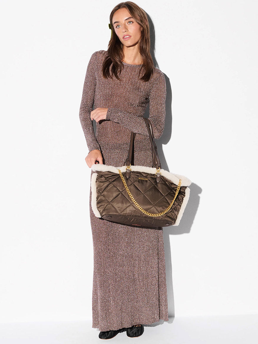 A woman in a long-sleeved, floor-length brown dress holds the MZ Wallace Medium Madison E-W Tote in Shearling & Walnut, featuring Italian leather trim, a gold chain handle, and quilted detailing.
