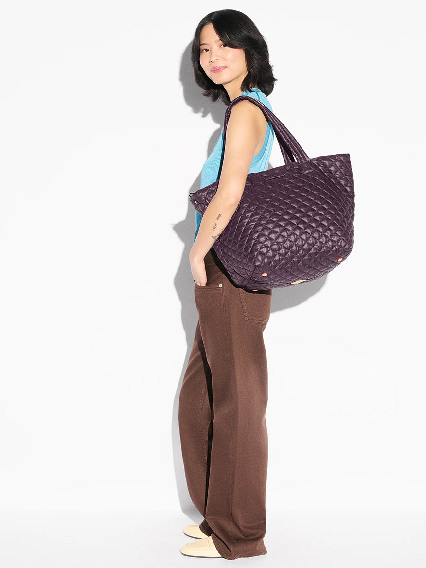 A young woman models the MZ Wallace Medium Metro Tote Deluxe in Raisin Oxford with brown wide-leg pants against a white background.
