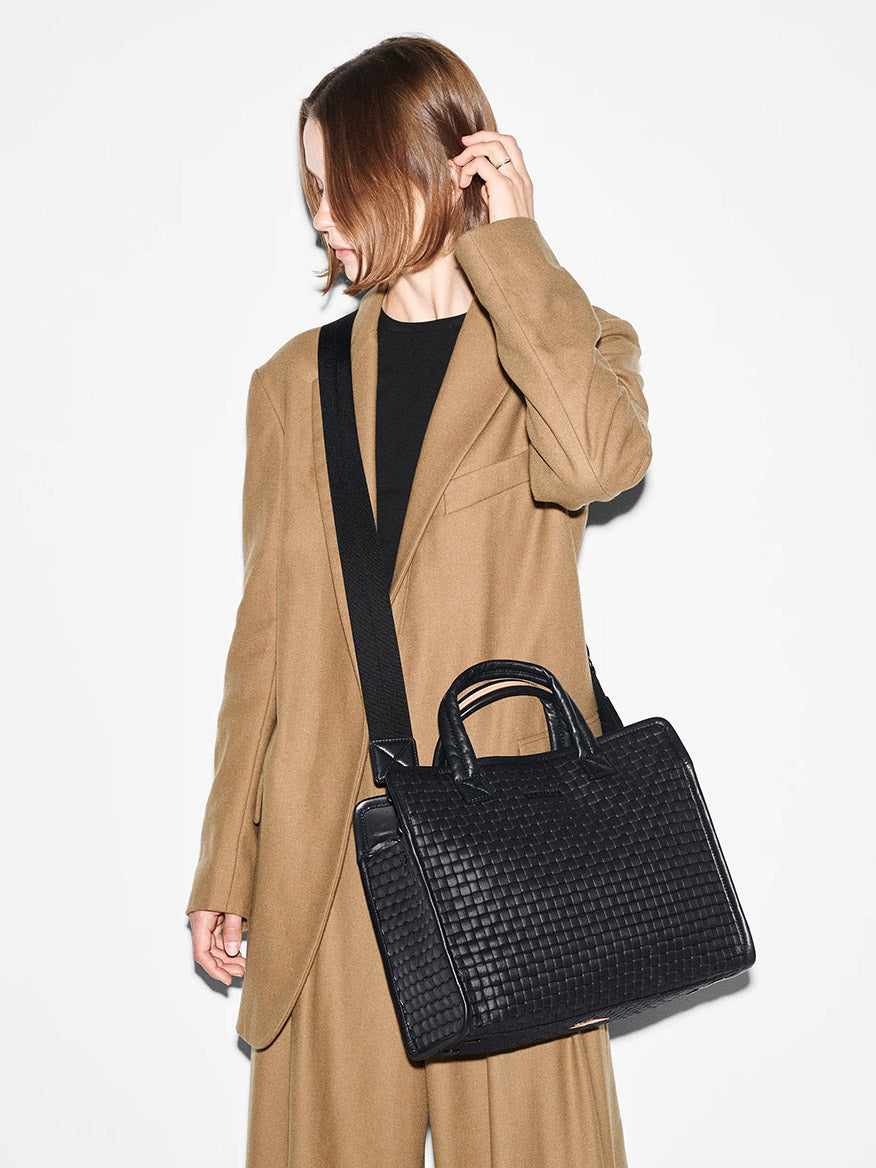 A woman in a stylish camel coat and black outfit holds a MZ Wallace Medium Woven Box Tote in Black Oxford, looking away from the camera, against a white background.