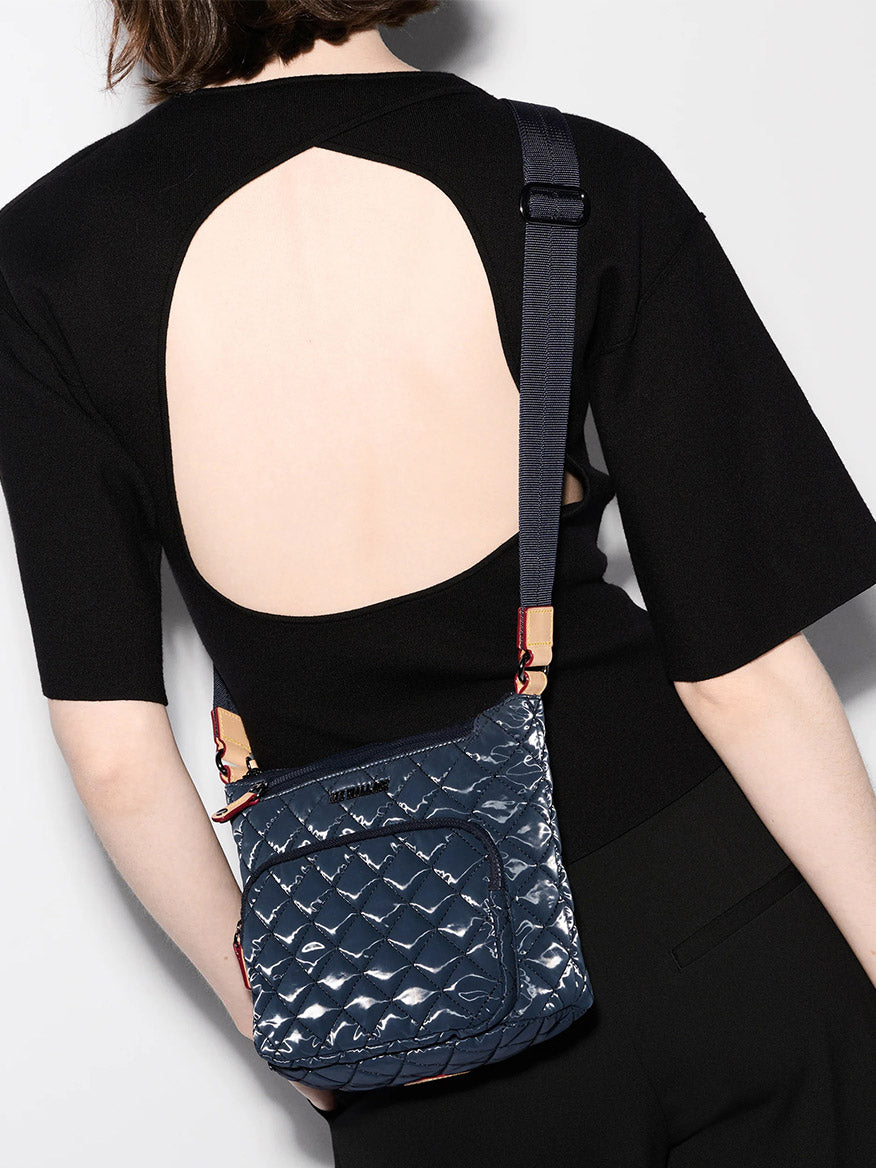 A person in a black open-back top carries the MZ Wallace Metro Scout Deluxe in Ink Liquid Oxford, a small, shiny crossbody bag made from quilted Italian leather in dark blue.