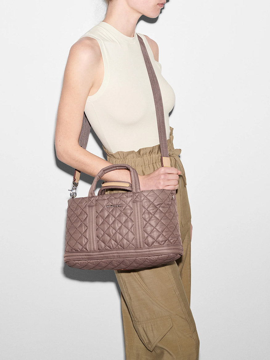 A woman in a sleeveless top and khaki trousers carries a MZ Wallace Metro Utility Crossbody in Mauve Oxford on her shoulder against a white background.
