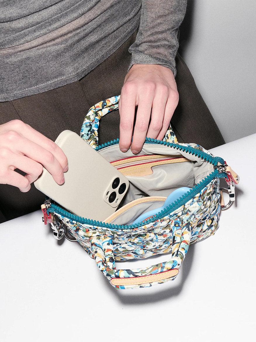 A person places a smartphone into an MZ Wallace Micro Sutton in Club Paint Oxford, its open zipper revealing organized inner compartments.